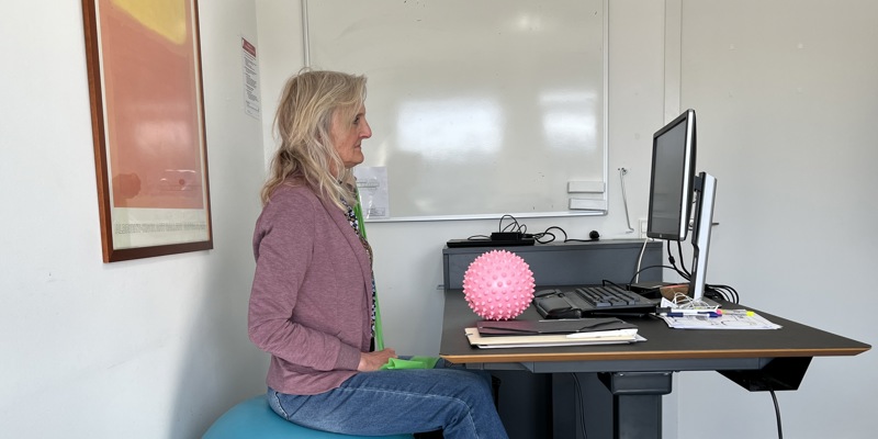Person sidder på yogabold ved skrivebord med computerudstyr.