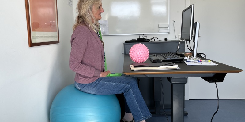 Person sidder på yogabold ved skrivebord med computerudstyr.